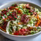 Colorful cabbage salad with sun-dried tomatoes, carrots, and herbs, tossed in a glass bowl with salad tongs.